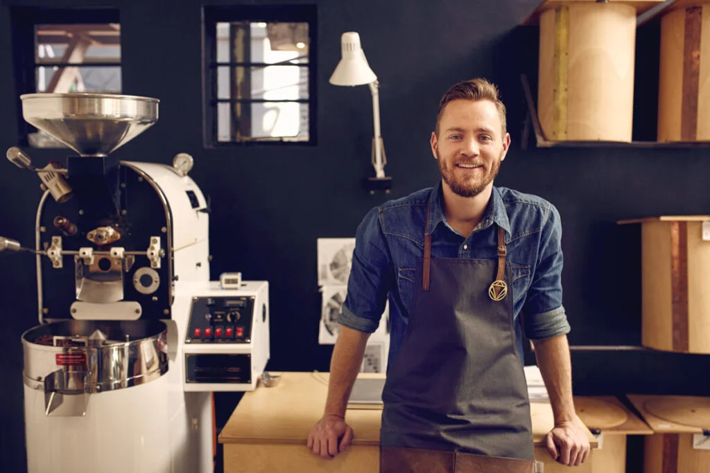 a man in a work apron smiling in front of a large coffee grinder