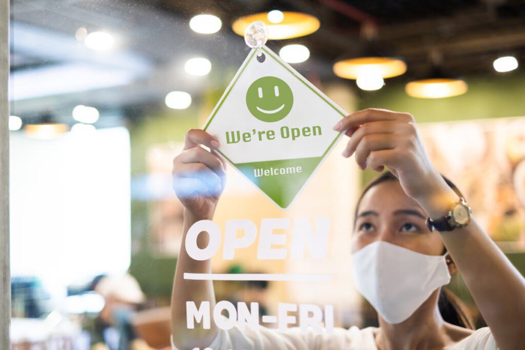 a woman in a mask puts up a sign that says "we're open, welcome"