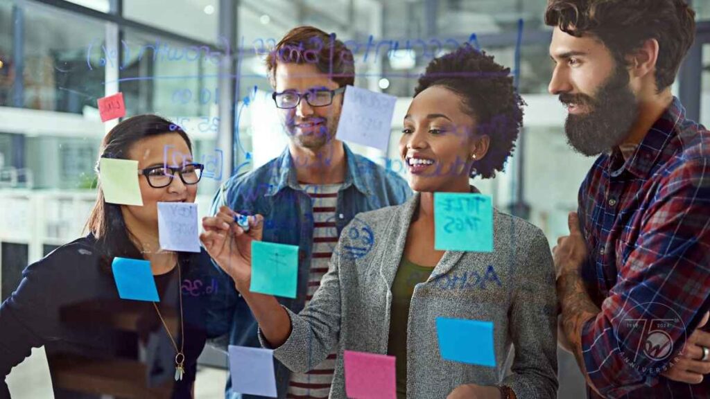 Four professionals collaborating at a brainstorming session, writing ideas on a clear board covered with sticky notes and handwritten notes, symbolizing teamwork and innovation in planning for nonprofit trends in 2025.
