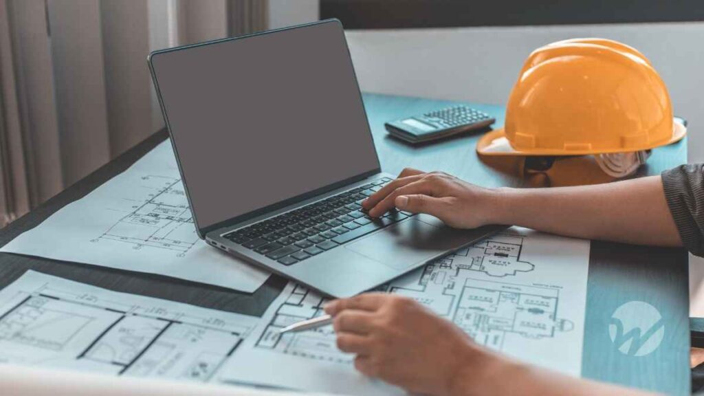 Construction professional working on laptop with architectural blueprints, hard hat, and calculator—symbolizing tax planning and compliance for the construction industry under new OBBBA legislation.