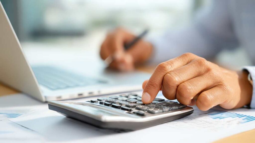 A person using a calculator at a desk with financial documents and a laptop, representing nonprofit accounting and capital campaign cost tracking.
