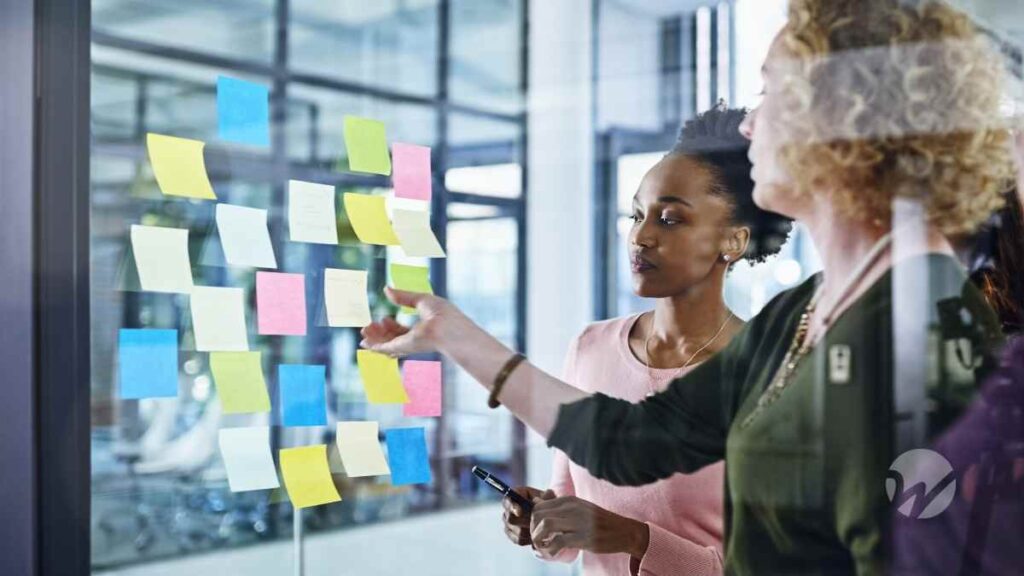 Two nonprofit professionals collaborate on planning and compliance tasks, organizing ideas on a wall of colorful sticky notes.