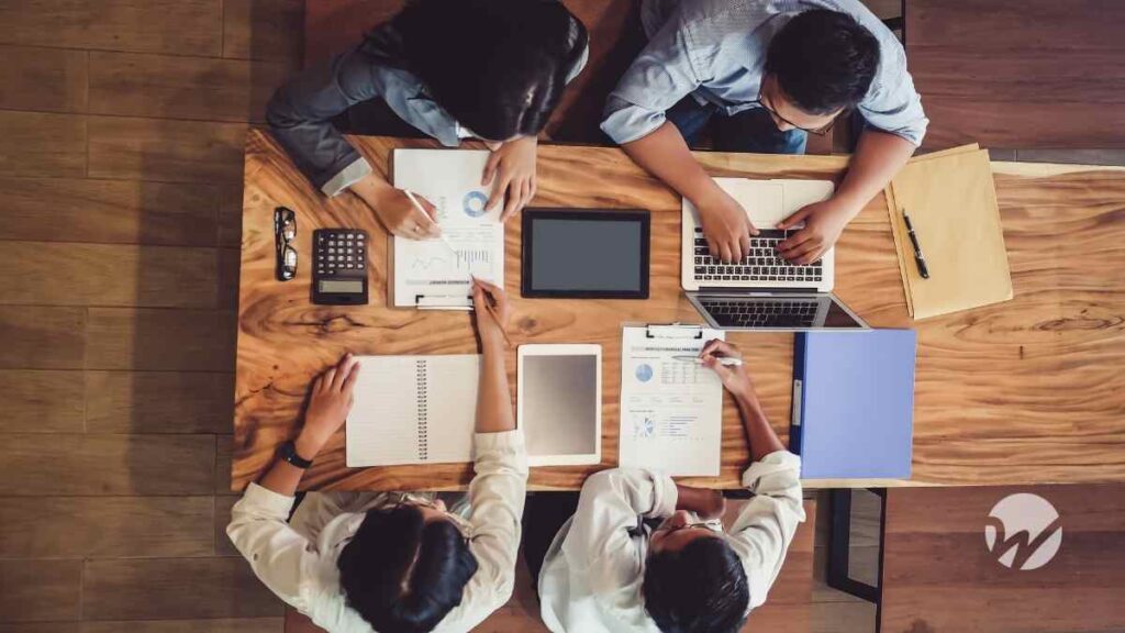 Overhead view of four professionals working together at a wooden table with laptops, financial reports, charts, and notebooks during a collaborative meeting.