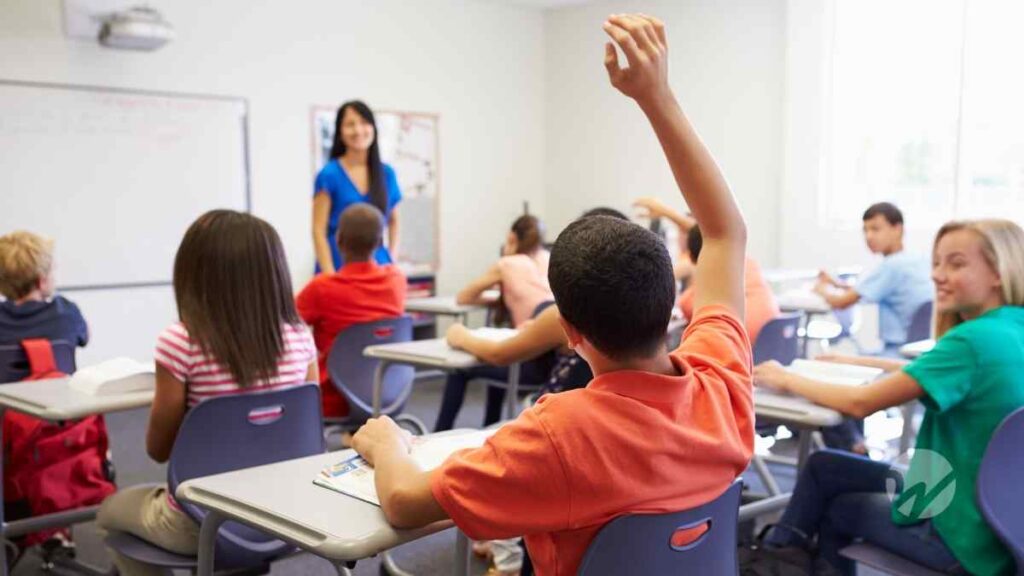 Child raising hand in classroom setting with teacher in front of white board
