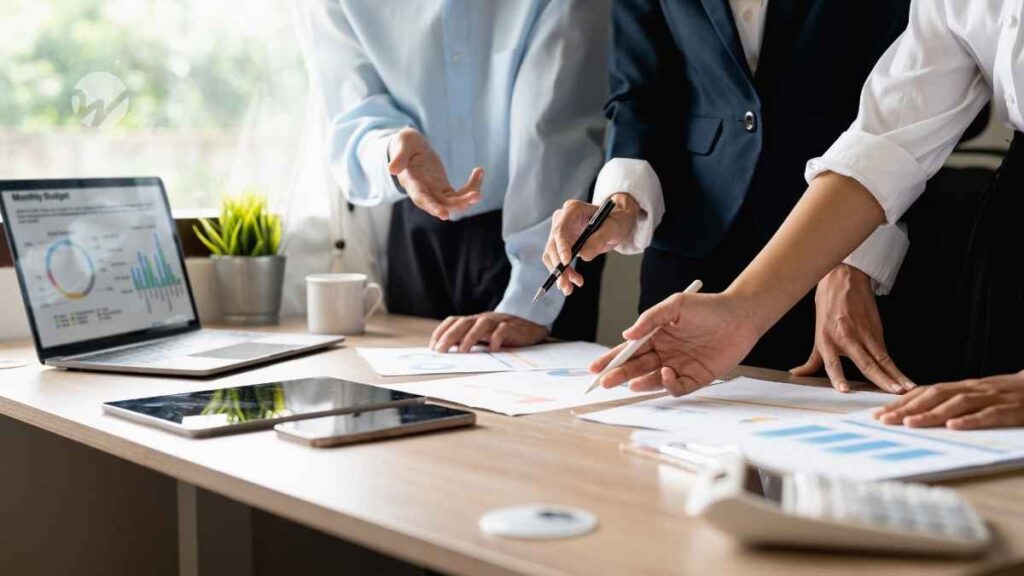 three professionals grouped around a desk looking at financial statements and dashboards, only arms and table visible