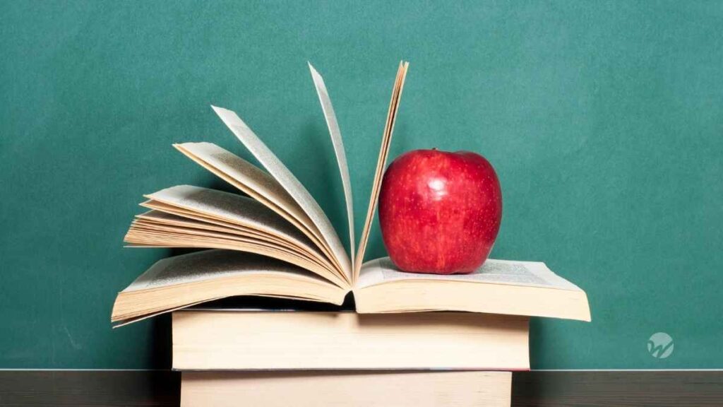 Stack of books in front of empty green chalkboard. Apple balance on the pages of an open book.
