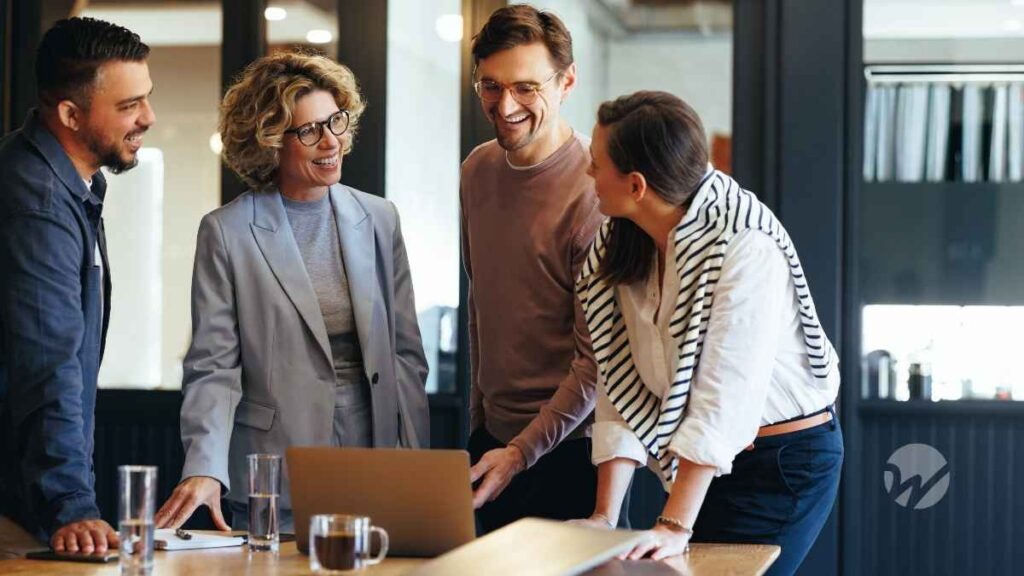 four casually dressed professionals stand around conference table over laptop, smiling