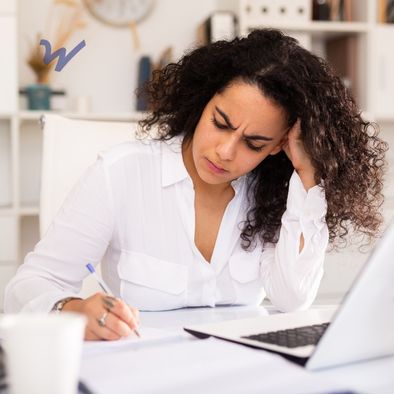 frustrated professionally dressed woman sits in front of laptop and writes on notepad
