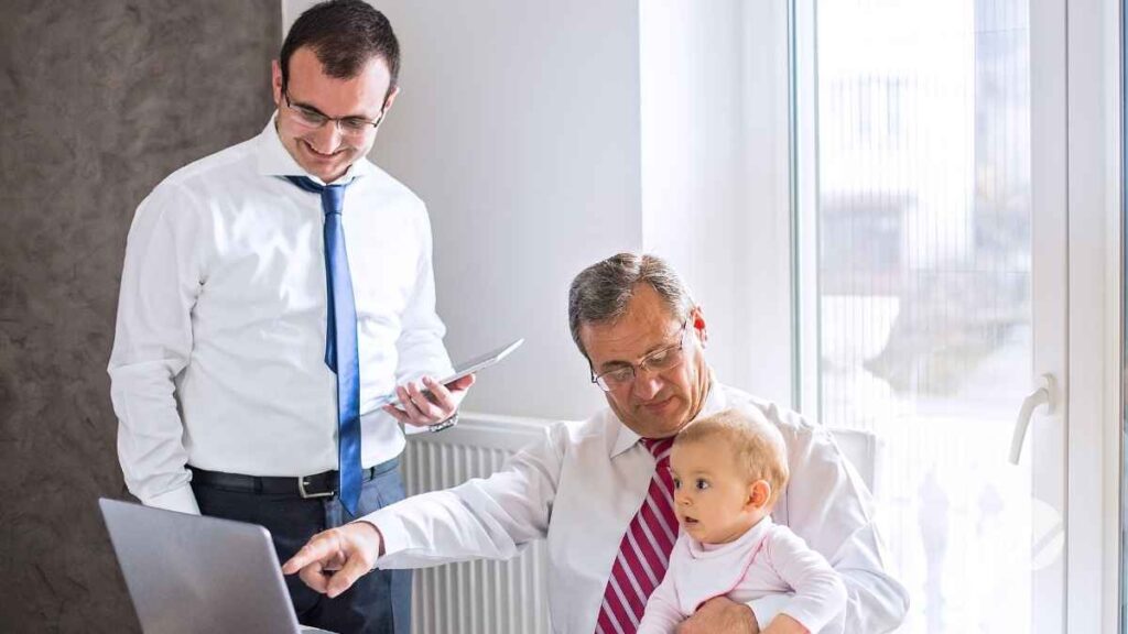 grandfather holds baby and points to laptop screen while standing next to baby's son and dad