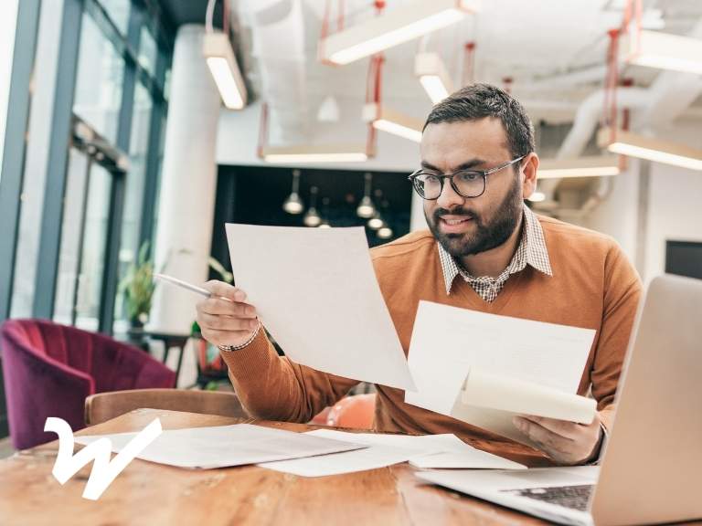 Man with beard and glasses evaluating several documents at table in front of laptop